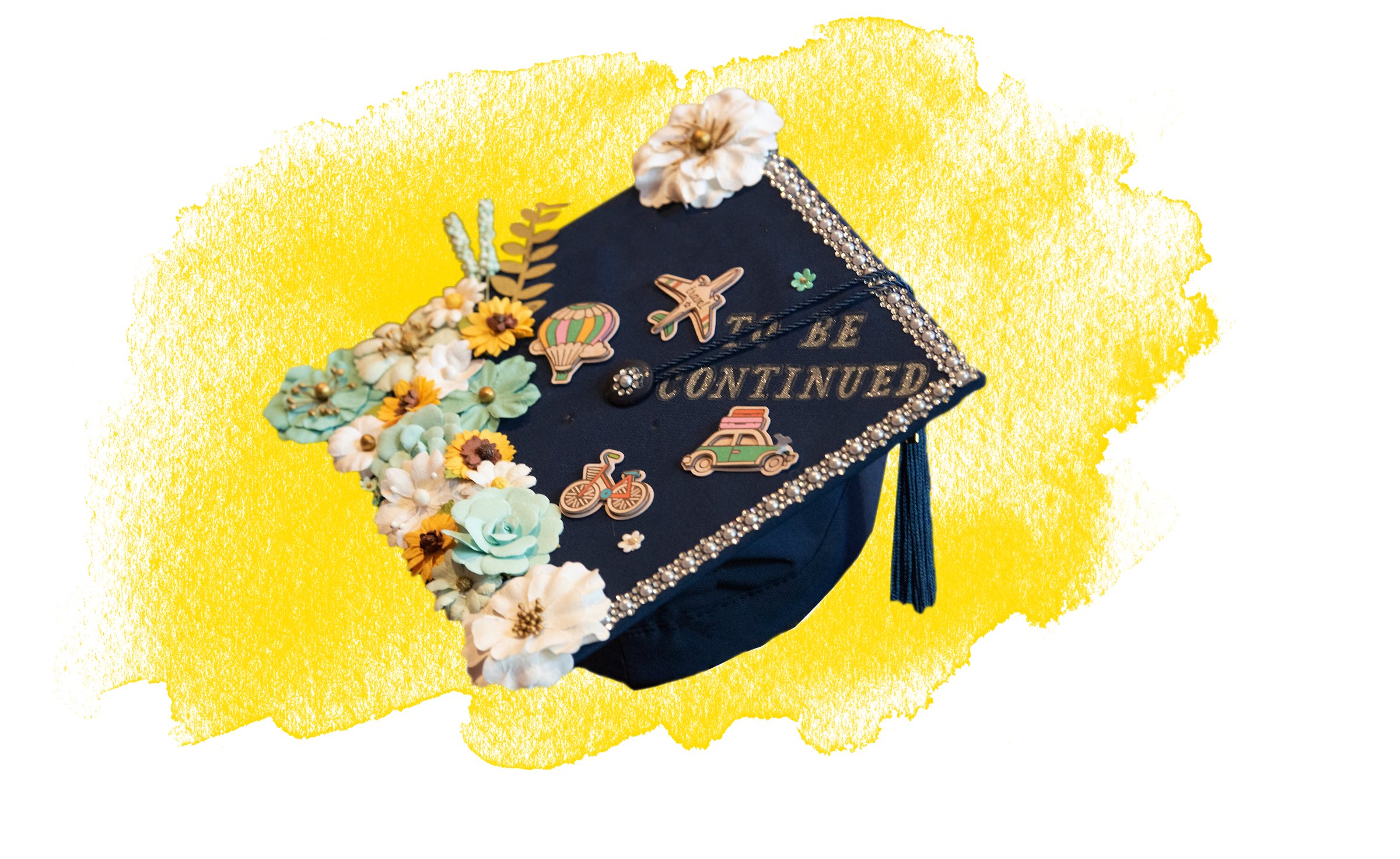 Yellow watercolor brushstroke in the background with a decorated grad cap with paper flowers, scrapbook decorations, and a tassle on top.