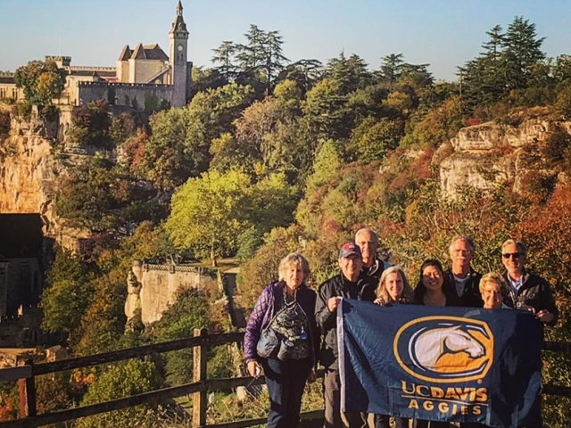 A group of Aggie Adventures travelers pose for a photo in Rocamadour, France. A hot-air balloon is visible in the background floating in the air.
