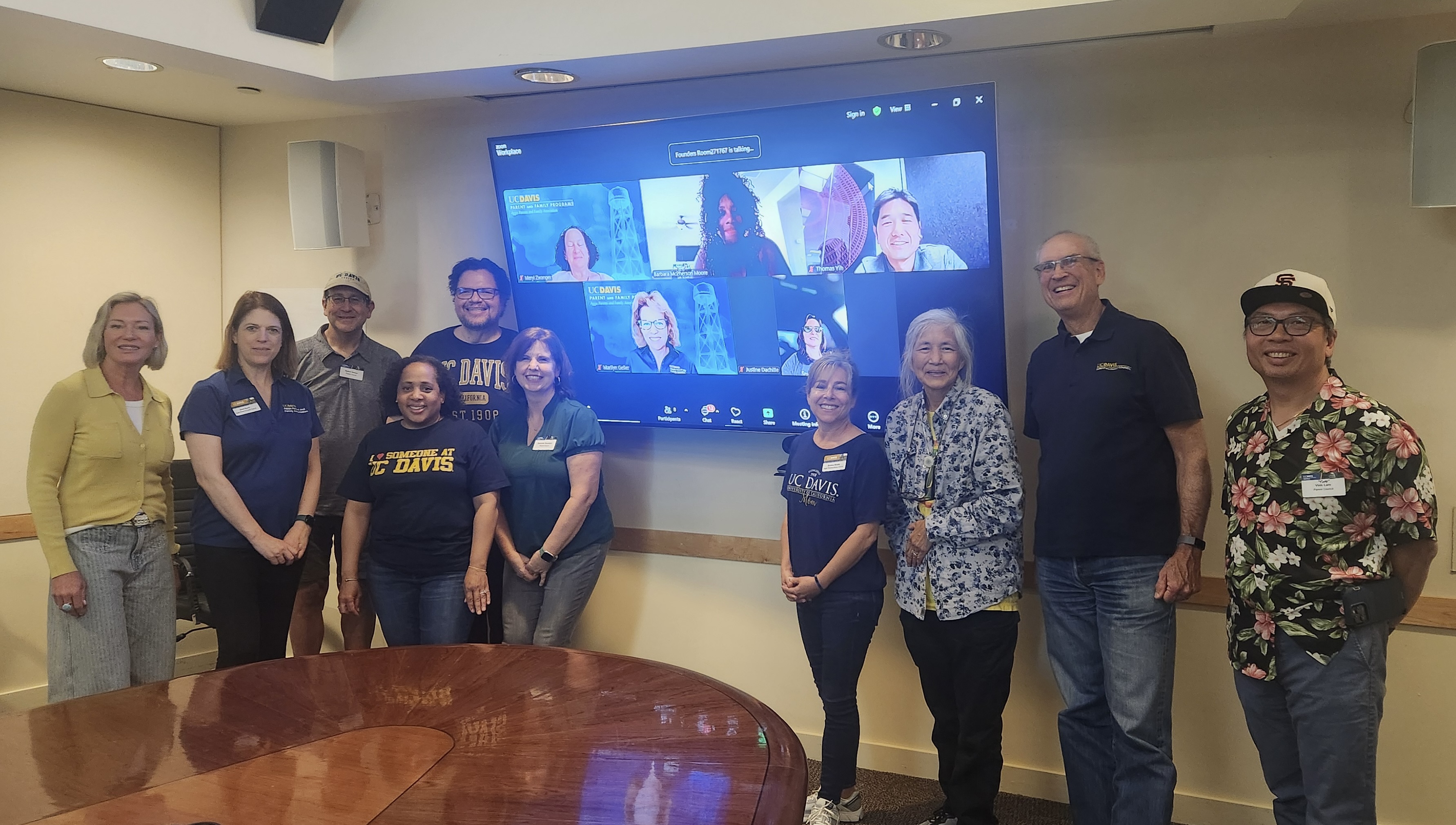 A group of people standing next to a tv with a zoom meeting on it