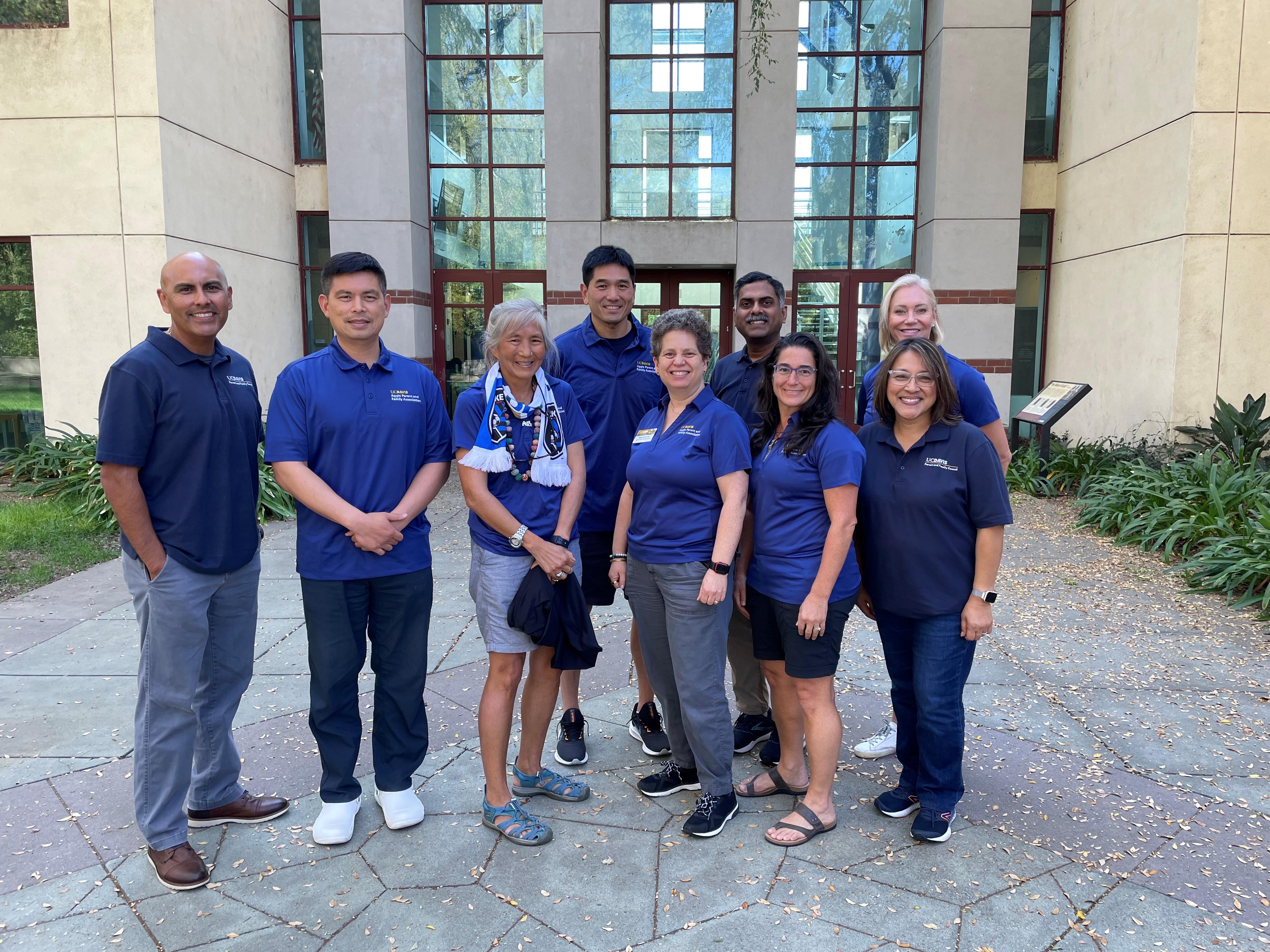 a group of parent council members stand in front of the alumni center at UC Davis. They are all wearing blue polos and smiling. 