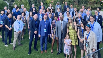 A group of UC Davis alumni, leadership, community members, and Bernadette Meehan pose for a photo on the grass. Hedges with pink and red flowers and trees can be seen behind them, as well as the cityscape and mountains.