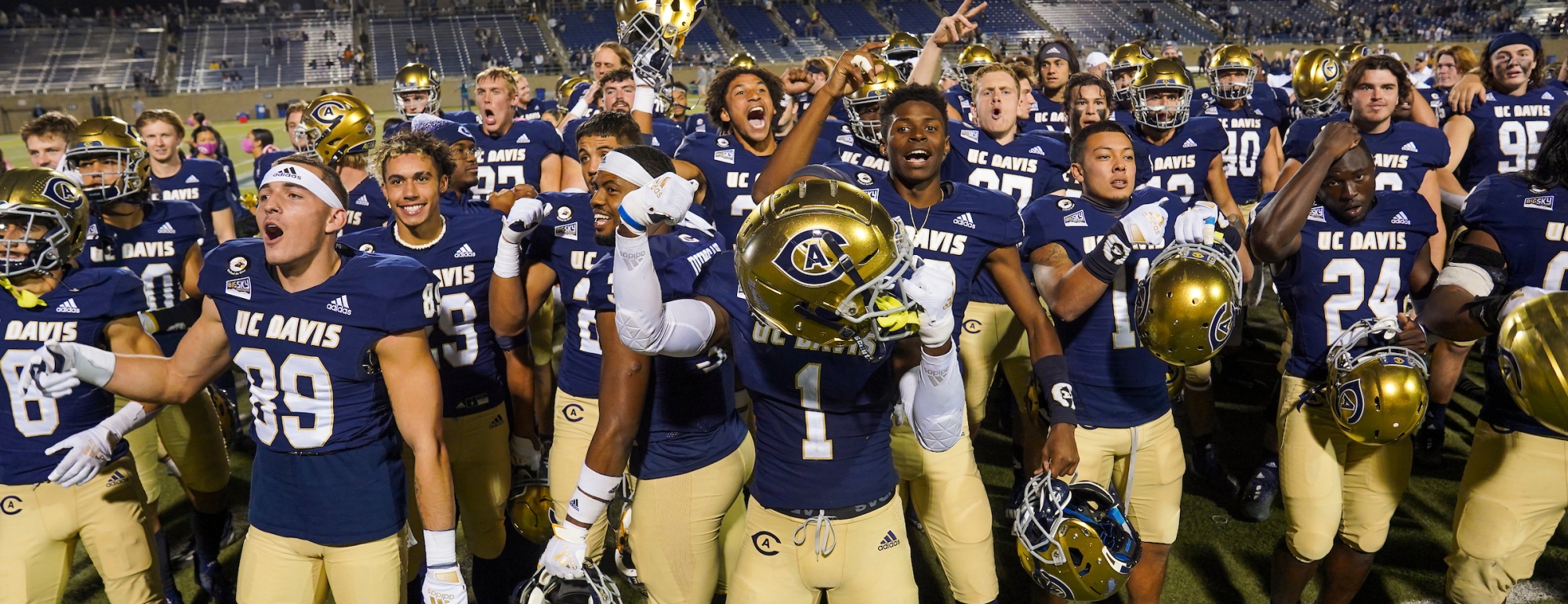 A group of UC Davis football players on the field with their hands in the air while holding onto their helmets.