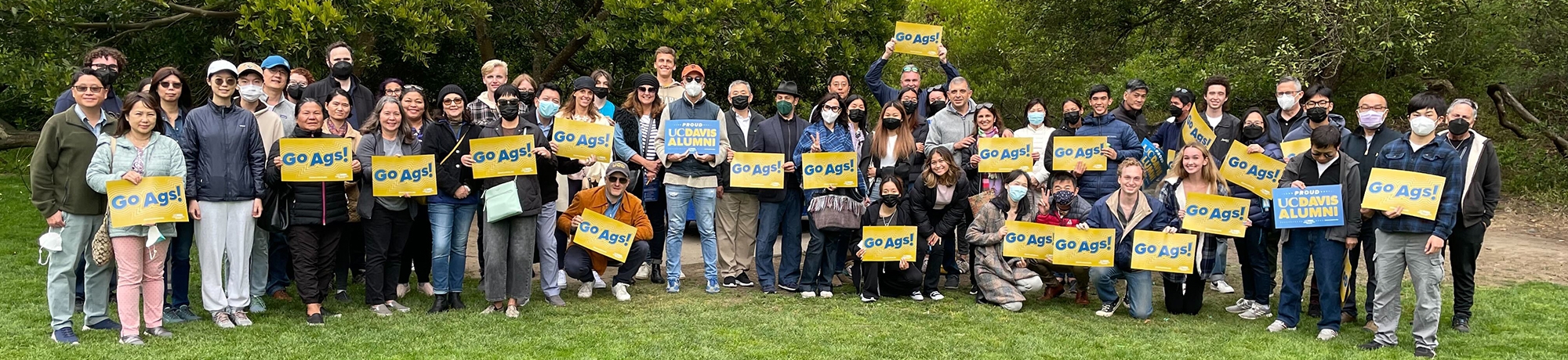 Variety of individuals wearing masks, standing in a group, and holding up multiple signs that say "Go Ags!" and one that says "UC Davis Alumni" 