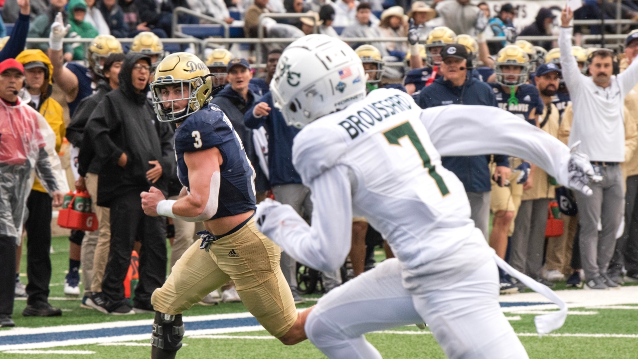 UC Davis football players runs with the ball with a Sac State player in the foreground chasing him