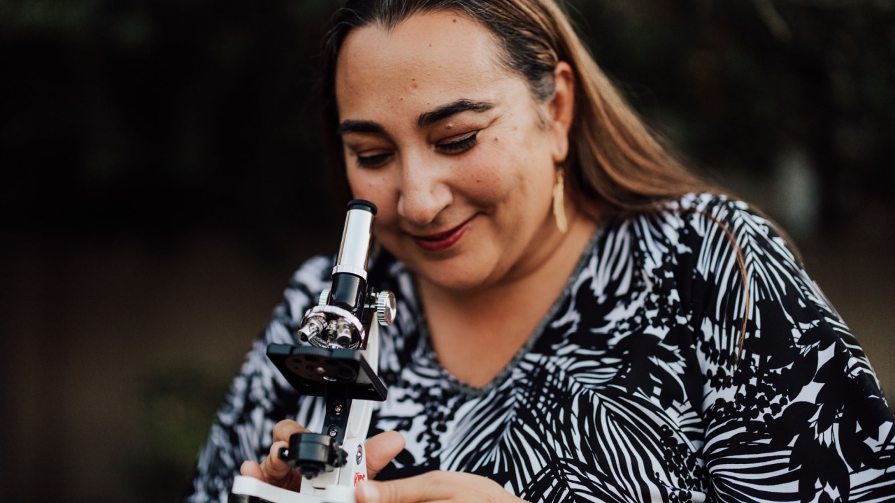 Nazzy, wearing a white top with black print patterns of palm leaves, poses with a white and black microscope in her hands while peering down at it. She is outdoors.