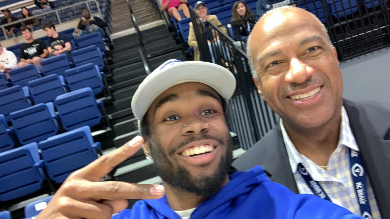 Two people pose for a selfie at a UC Davis sporting event. One person is wearing a white ballcap and blue sweatshirt and the other is wearing a gray suit with a blue and yellow checkered shirt.