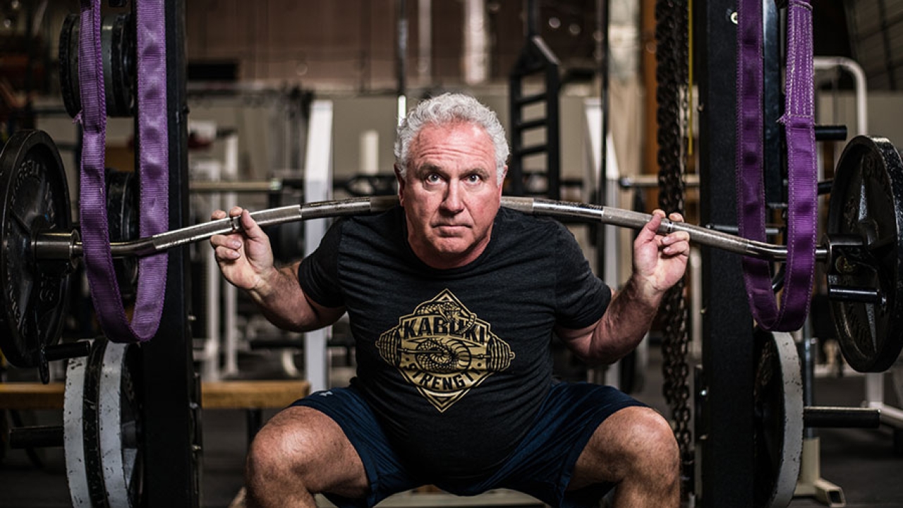 Rudy Kadlub squatting and holding a barbell at his shoulder in a gym.