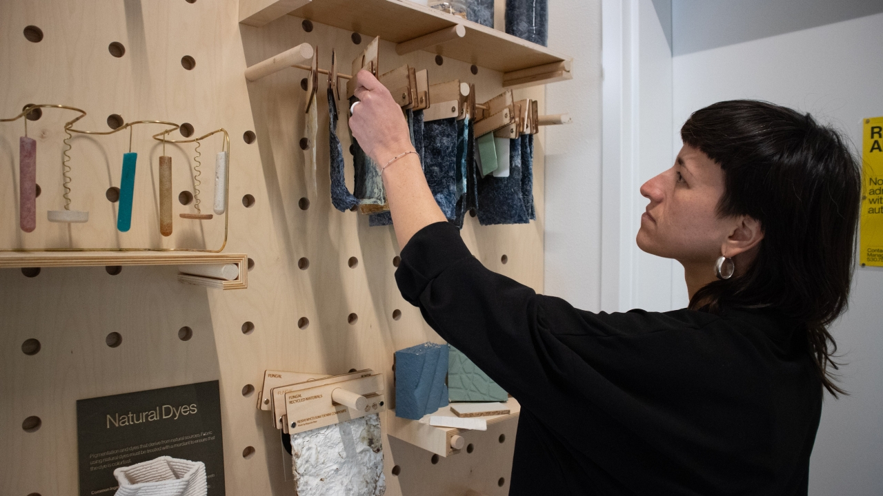 A person inspects a fabric sample hanging off a wooden peg on a craft board mounted on the wall.