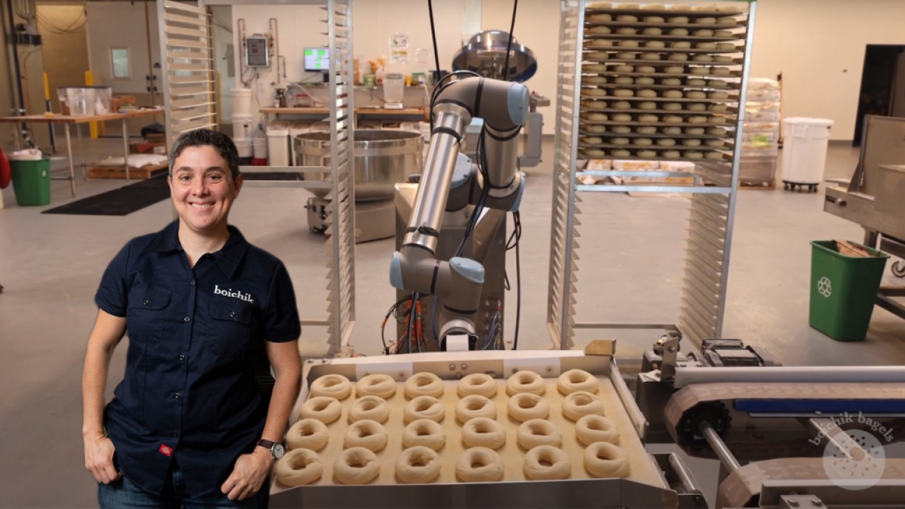 Emily Winston wearing a "Boichik Bagels" button-down shirt placed in front of an assembly line with a bagel-making robot and a tray of unbaked bagels.