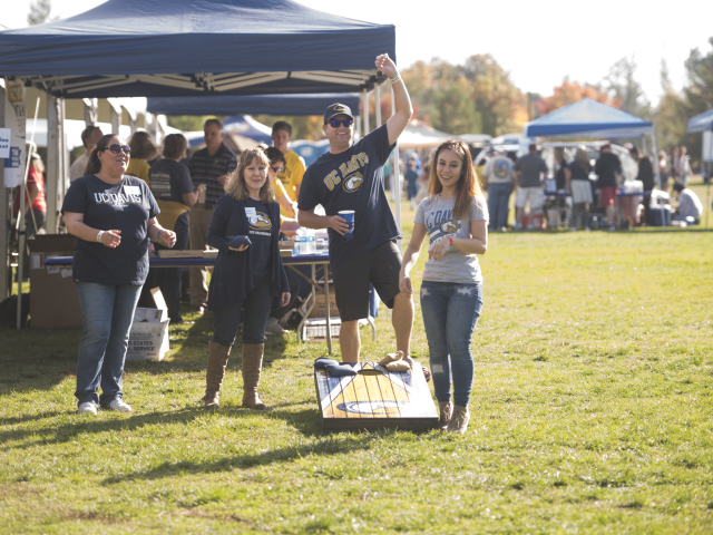 A group of UC Davis family members wearing "UC Davis" apparel play cornhole on the lawn in front of a canopy.