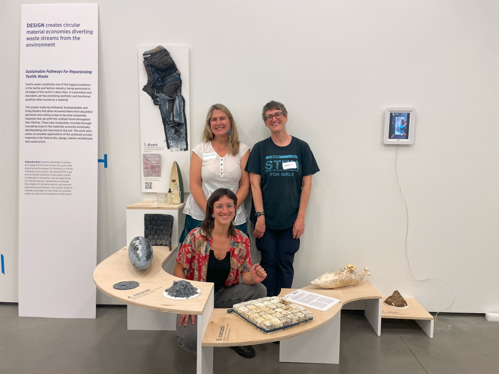 A group of three people pose behind a display showcasing different types of textile waste laid out on multiple platforms.