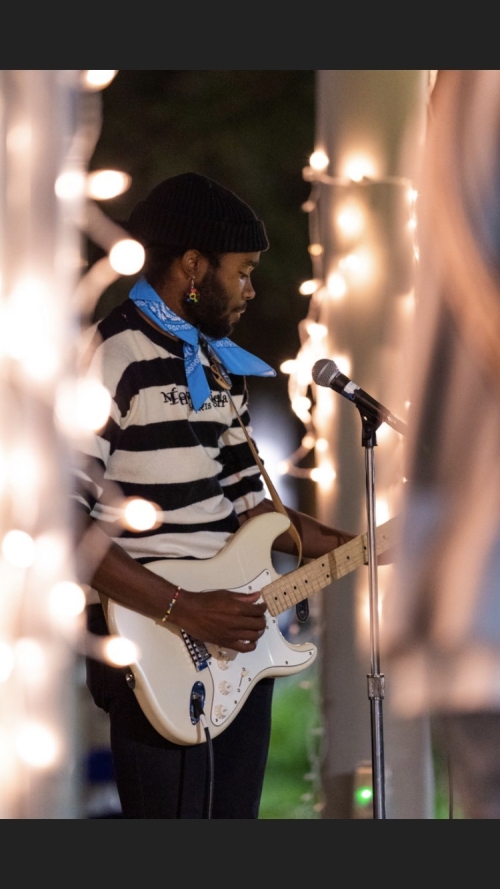 A person wearing black and white striped shirt with a blue bandana around their neck is playing the guitar and singing on stage.