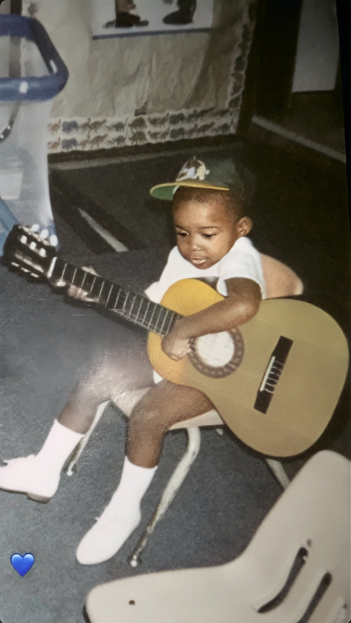 A young child wearing an Oakland A’s baseball cap, white shirt and white socks and shoes sits on a chair and strums a guitar.