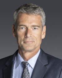 Older male poses for a headshot indoors wearing a dark grey suit, light blue button-down and dark tie.