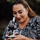 Nazzy, wearing a white top with black print patterns of palm leaves, poses with a white and black microscope in her hands while peering down at it. She is outdoors.