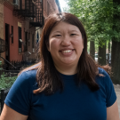 Bessie wearing a blue T-shirt stands in front of brownstone apartments and trees.