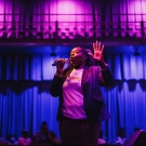 Bland holding a microphone and animatedly reciting poetry, holding one hand up, in a dark theater with blue and purple lights in the background