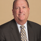 Middle-aged male poses for a headshot wearing a grey suit, white and light blue checkered button-down shirt, and gold and blue tie.