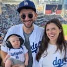 Sam Fleischer stands with wife and baby at Dodgers stadium. Each are wearing white and blue Dodgers jerseys. 