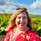 Ginger Welsh wearing sunglasses on her head and a red top with white flowers in front of a vineyard with blue skies.