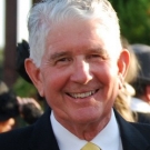 Older male poses for a photo outdoors wearing a white button-down, gold tie, and dark blazer.