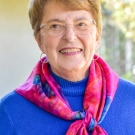 Older female poses outdoors wearing a blue sweater, fuchsia scarf, glasses, and gold hoop earrings.