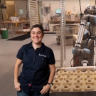 Emily Winston wearing a &#34;Boichik Bagels&#34; button-down shirt placed in front of an assembly line with a bagel-making robot and a tray of unbaked bagels.