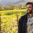 Derek poses with a black overcoat and grey button-down shirt. Vineyards and hills are visible in the background.
