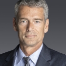 Older male poses for a headshot indoors wearing a dark grey suit, light blue button-down and dark tie.