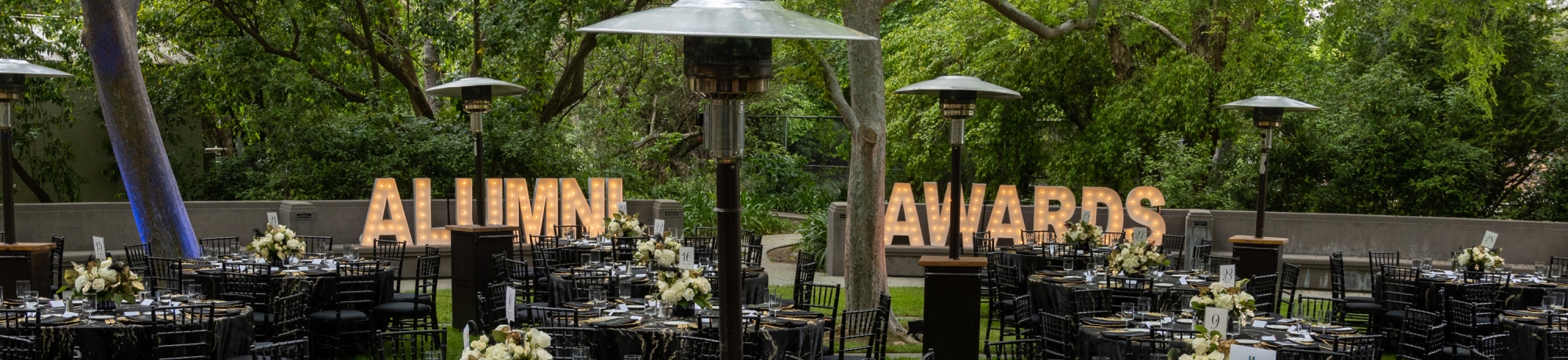 Round banquet tables on the grass with two marquee signs lit up that spell out the words "Alumni Awards"