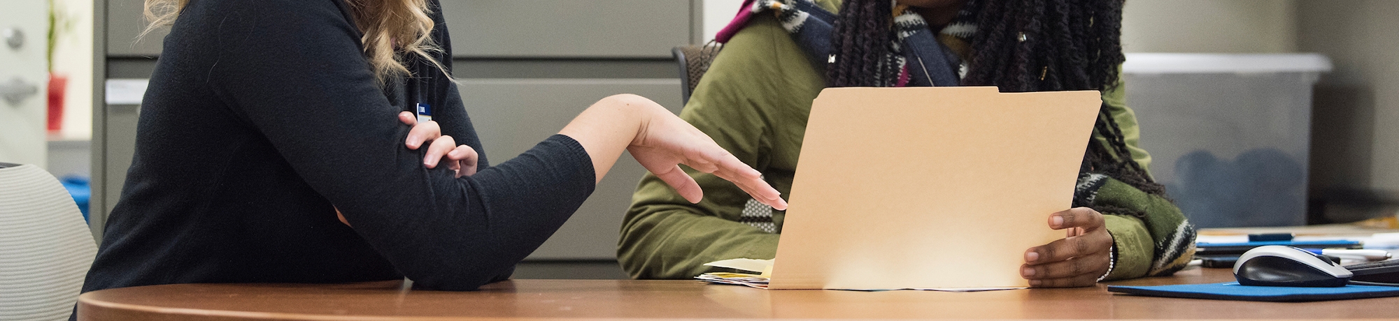 Two people having a conversation at a desk with a manila folder open.