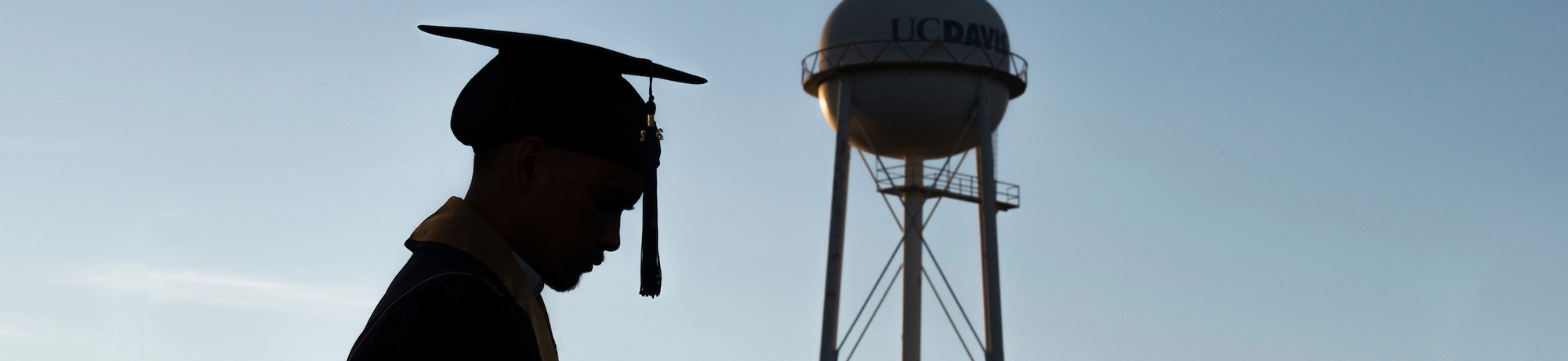 Silhouette of graduate in regalia with blue sky and UC Davis water tower in the background.