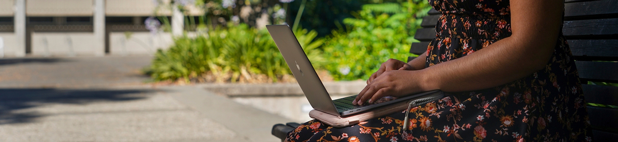 Person in floral dress working on the laptop outside on a bench.